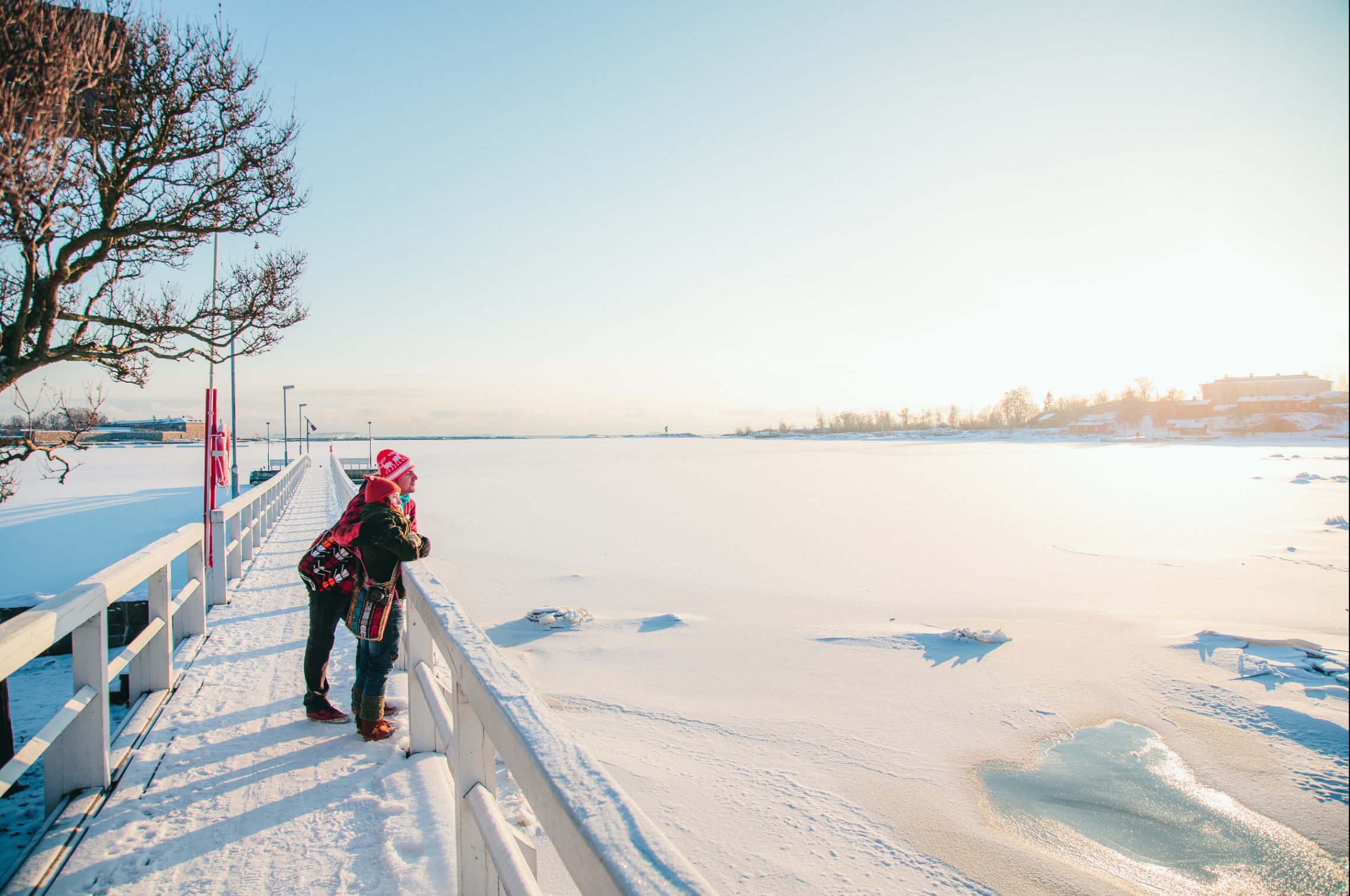 Kuvassa kaksi henkilöä seisoo valkoisella sillalla talvisessa ja aurinkoisessa säässä.