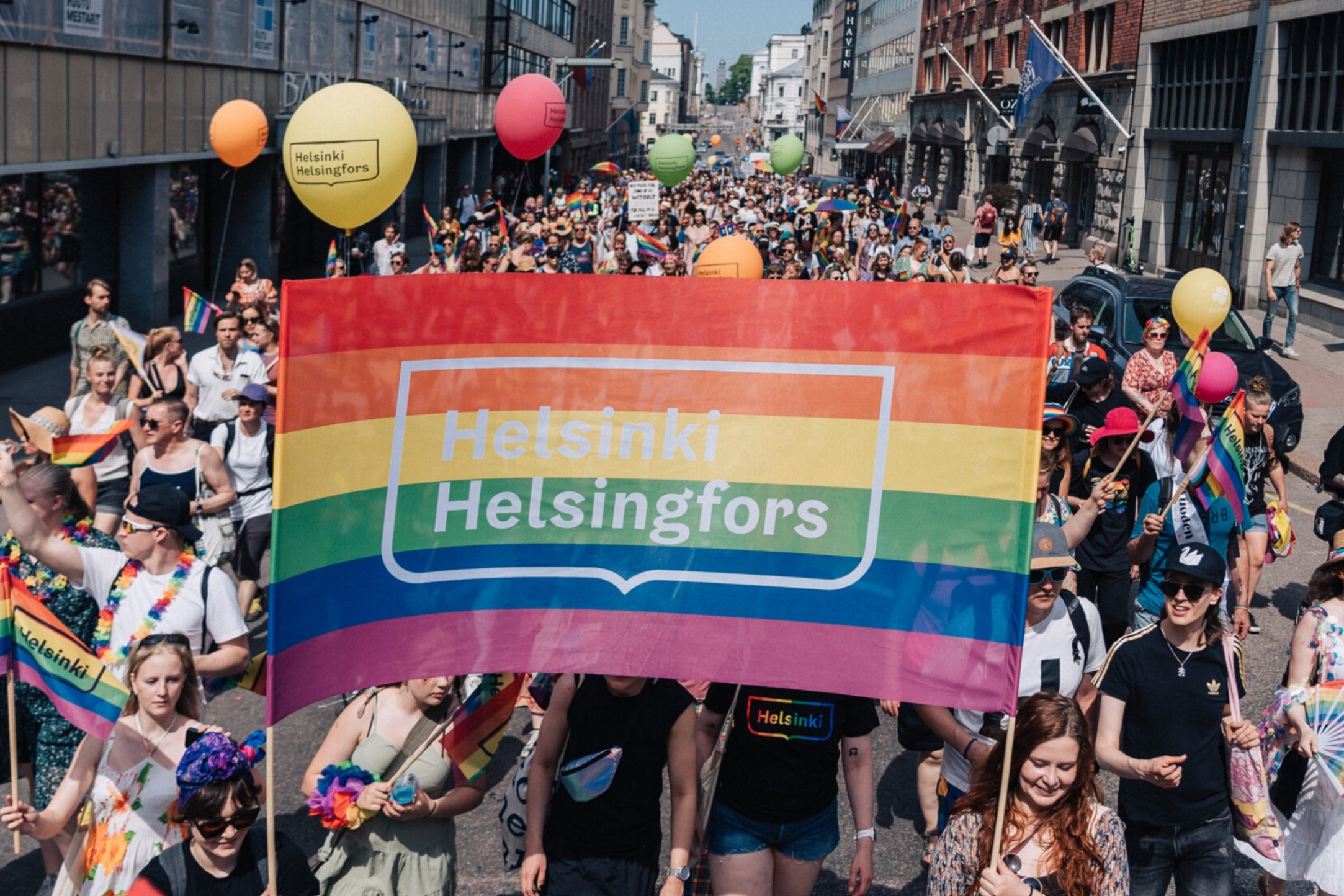 Kuva Pride-kulkueesta. Kuvassa etualalla sateenkaarilippu, jonka keskellä on Helsingin kaupungin logo valkoisella.