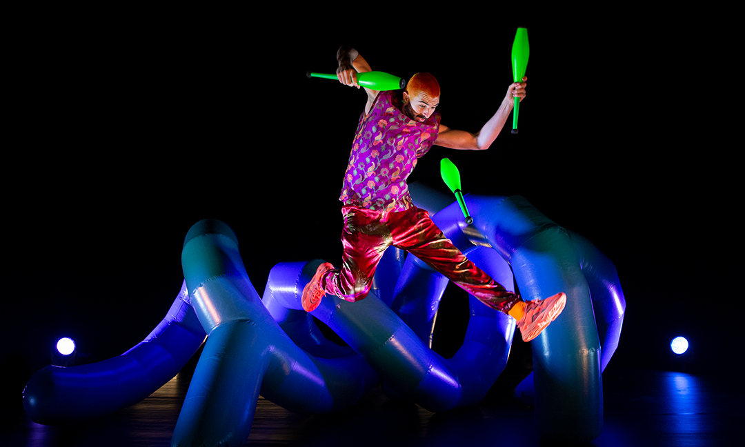 Kuvassa henkilö hyppää ja jongleeraa neonvihreillä keiloilla. Takana sininen ilmatäytteinen muovityynyke.