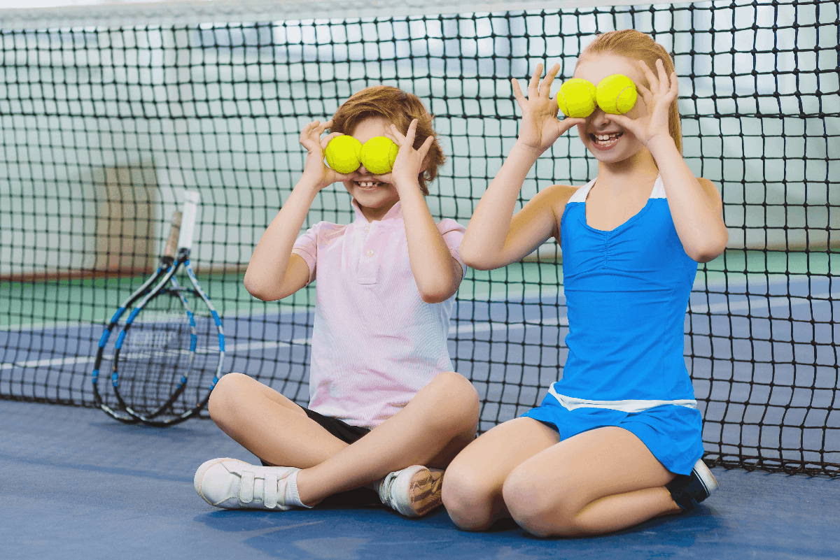 Kaksi lasta istuu tenniskentällä ja pitävät tennispalloja silmiensä edessä.