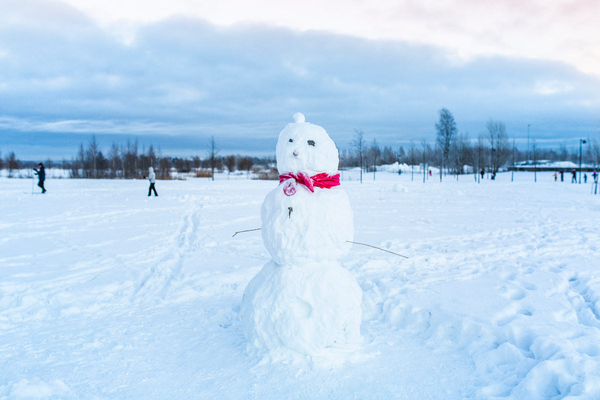 Lumiukko lumisessa ympäristössä. Lumiukolla on värikäs kaulaliina kaulassa. Taustalla kaksi hiitäjää.