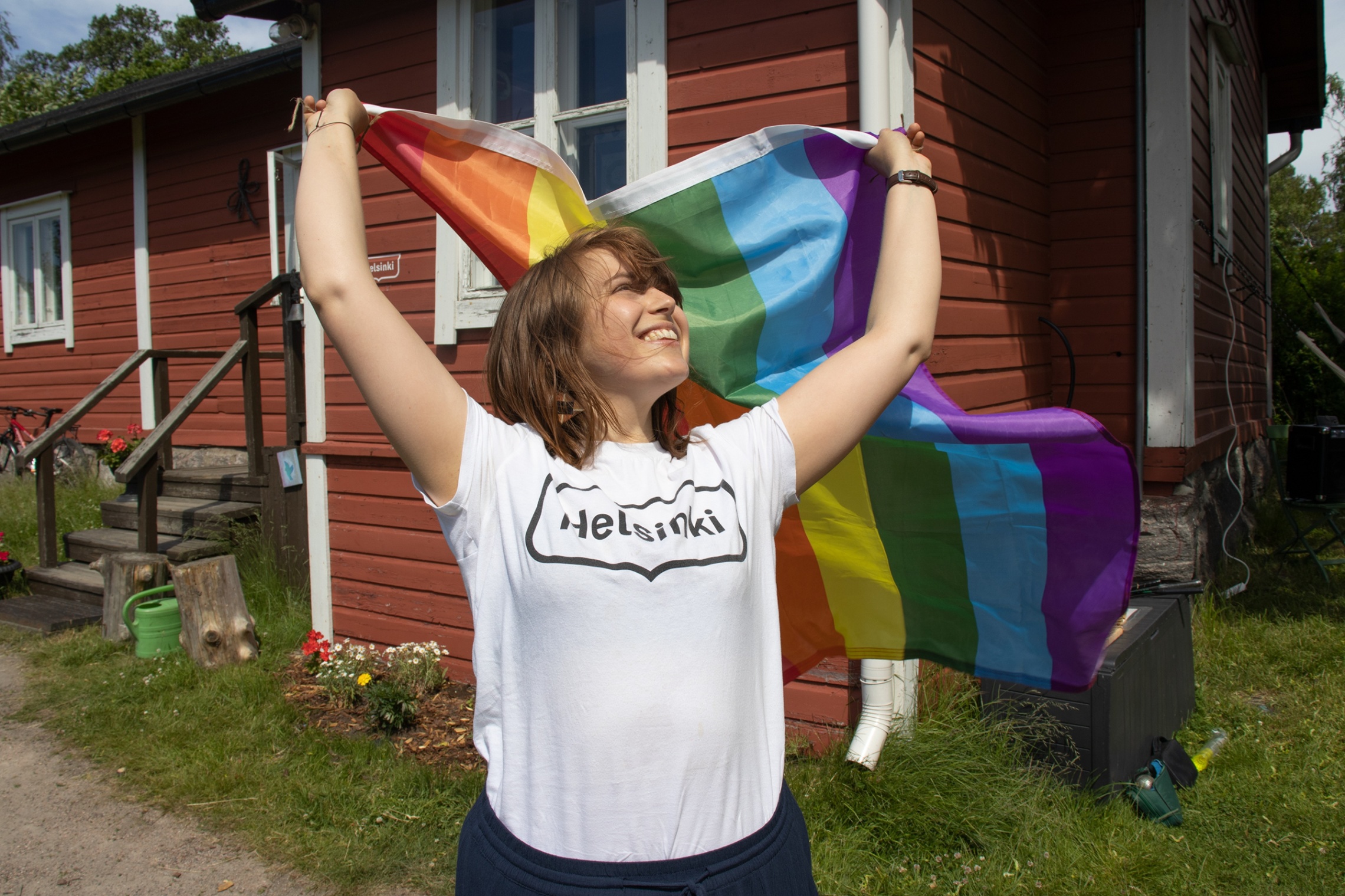kuvassa ihminen pitelee seisoo kädet ilmassa ja käsissään hänellä on sateenkaaren värinen pride-lippu. ihminen katsoo ylöspäin kohti lippua ja hymyilee