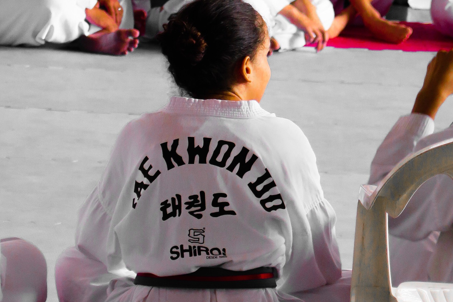 Henkilöt istuvat taekwondopuvussa liikuntasalin lattialla. Kuvassa yhden henkilön selkä kuvattuna lähempää ja puvussa näkyy erilaisia kirjaimia.