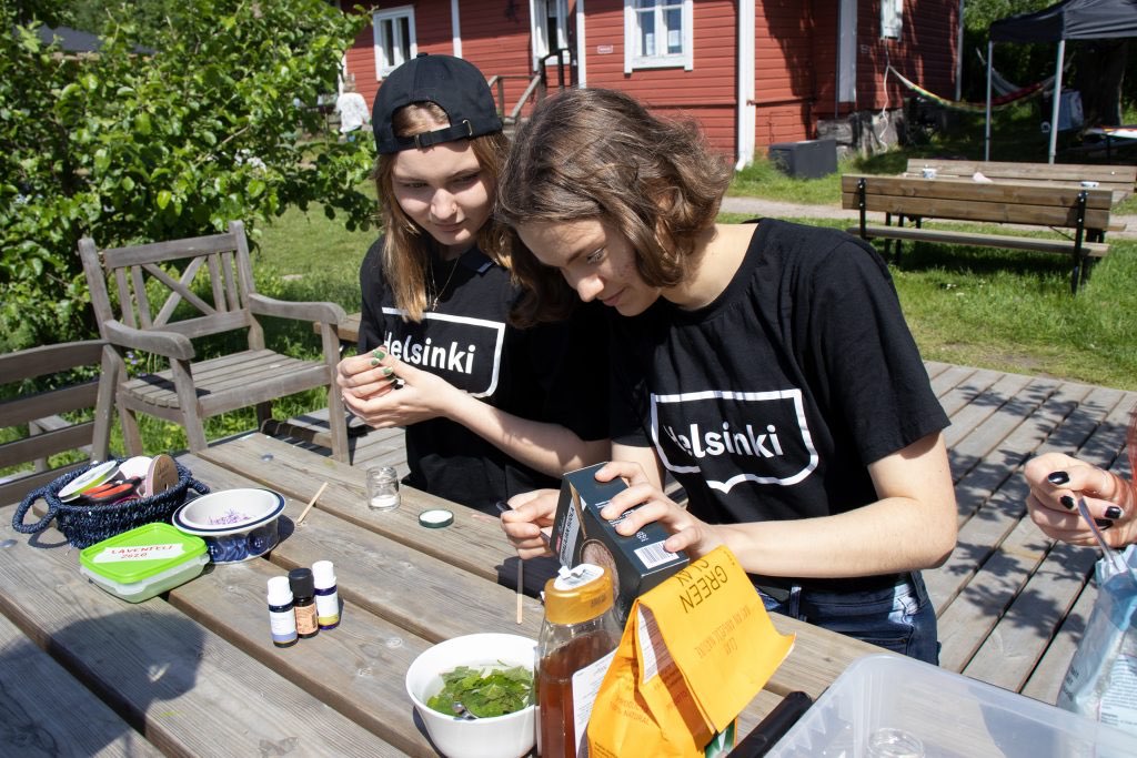 Kaksi nuorta Syötävässä puistossa Helsinki t-paidat päällä valmistavat luonnonkosmetiikkaa.