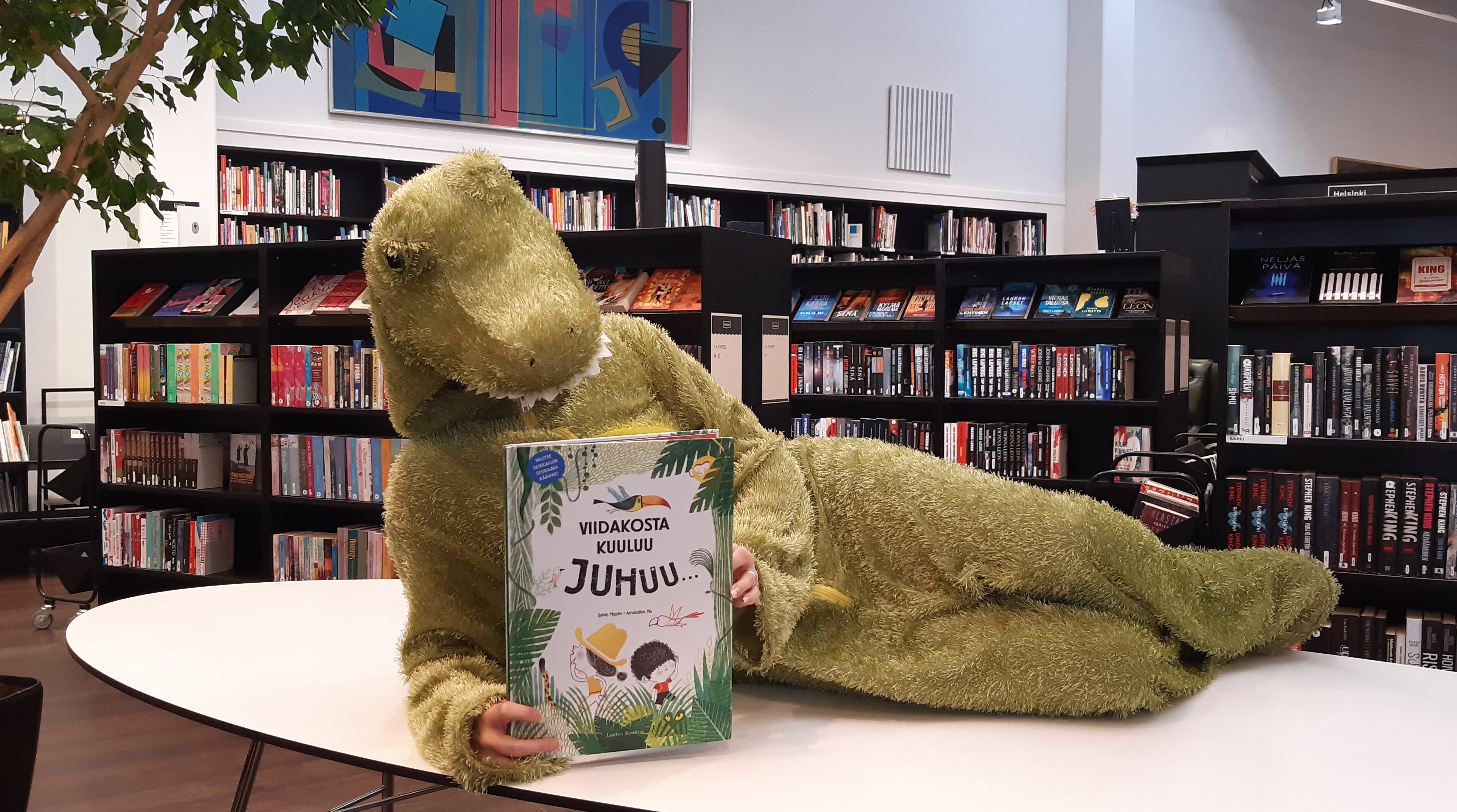 Krokotiiliasuun pukeutunut henkilö lukee kirjaa kirjastossa. Kuva Suvi Immonen
