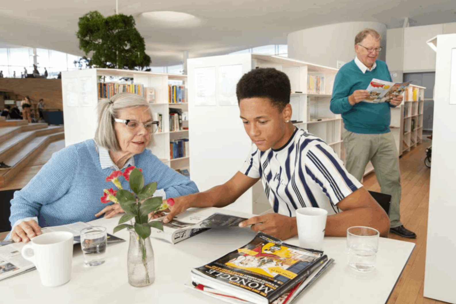 Seniori nainen ja nuori mies pöydän äärellä lukemassa samaa lehteä